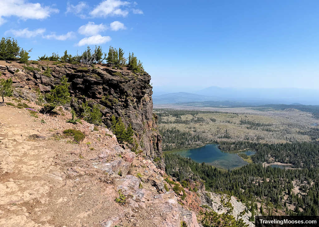Sheer rocky cliff edge along Tam McArthur Rim overlooking Little Three Creek Lake and pine forest below