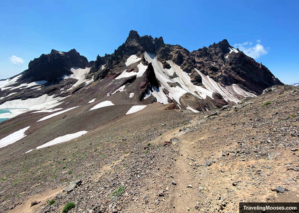 Winding dirt hiking path along an open scree ridge facing the dramatic, jagged spires and snow-filled chutes of Broken Top