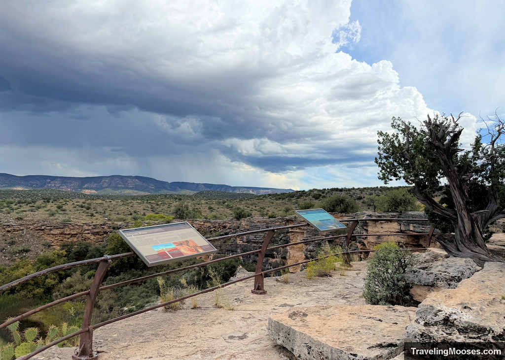 Metal safety railings and interpretive signs along the limestone rim trail at Montezuma Well with dark storm clouds gathering over the desert mountains.