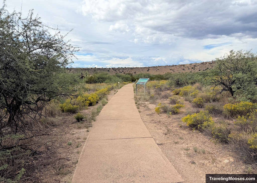 Paved upper loop trail lined with desert brush and interpretive signs under an overcast sky at Montezuma Well.