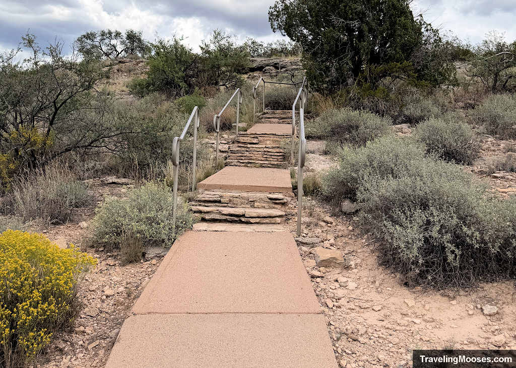 Paved trail and stone steps with metal handrails climbing through desert brush toward the Montezuma Well rim overlook.