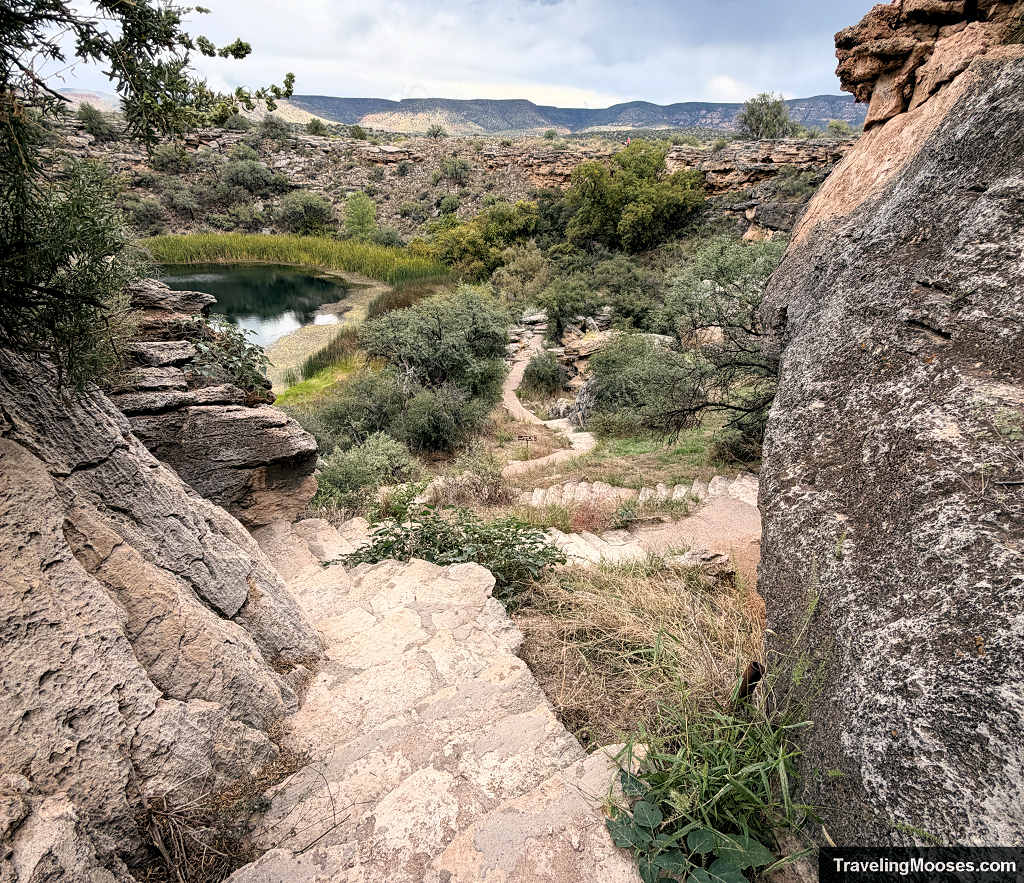 First-person view looking down a steep, native stone staircase descending through massive limestone rock formations into the Montezuma Well basin.