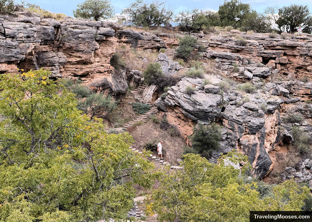 Narrow stone staircase winding down sheer limestone cliffs into Montezuma Well basin with a hiker descending toward the water.