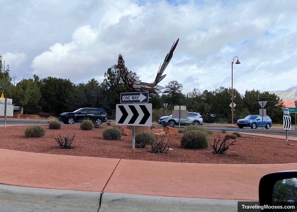 View from a driver seat waiting at a busy Sedona multi-lane traffic roundabout featuring a giant roadrunner sculpture in the median.