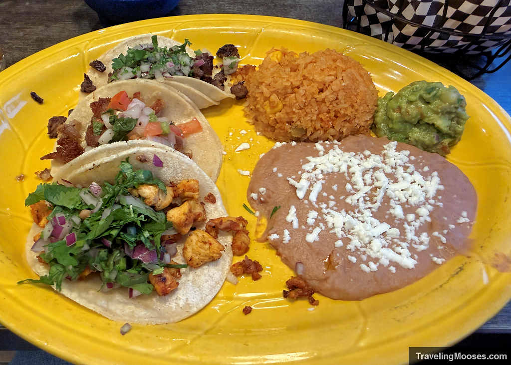 A yellow plate with three street tacos, Mexican rice, refried beans topped with cheese, and guacamole at a Sedona restaurant.
