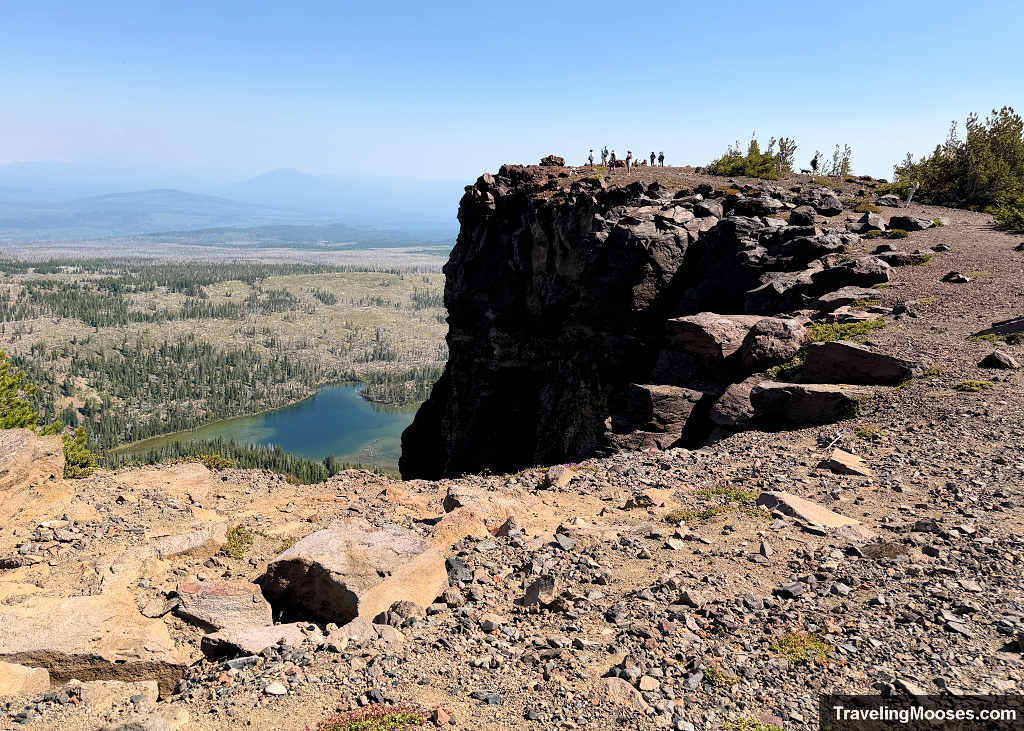 Rocky drop-off along Tam McArthur Rim looking across at hikers standing on the cliff edge with Little Three Creek Lake far below