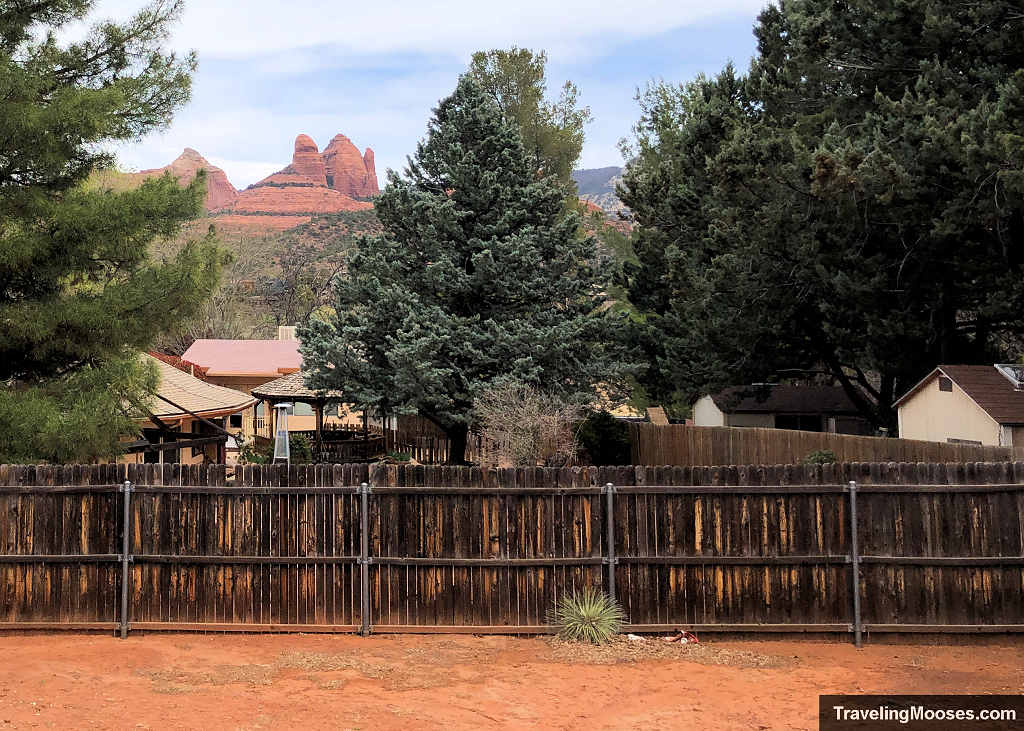 View from a West Sedona backyard fence looking toward evergreen trees and red sandstone formations in the distance.