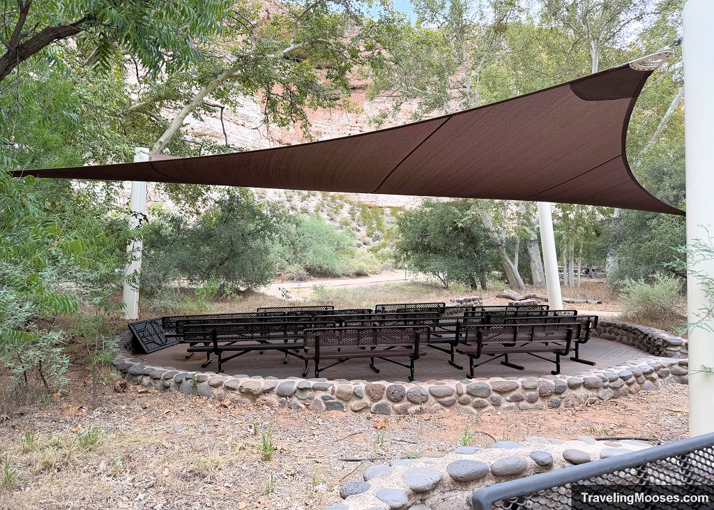 Shaded outdoor amphitheater seating area with benches under a large brown fabric shade sail and white support pillars