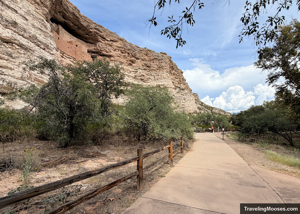 Paved accessible loop trail curving past a split-rail wooden fence with a distant view of Montezuma Castle in the limestone cliff wall