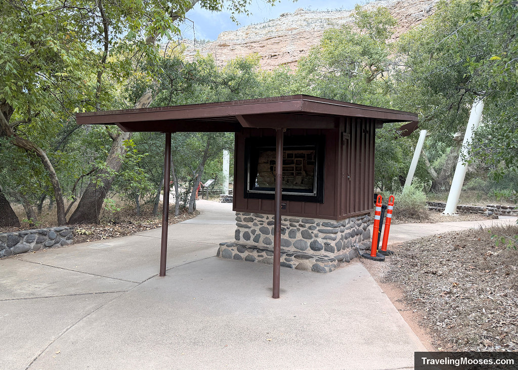 Brown wooden shelter kiosk with stone base housing an illustrated cutaway display along the paved walking loop