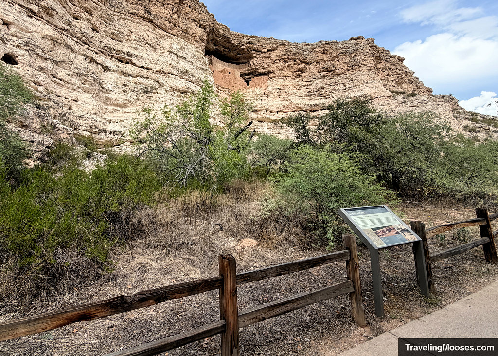 Trailside metal interpretive informational sign along the split-rail fence overlooking the cliff face and desert vegetation