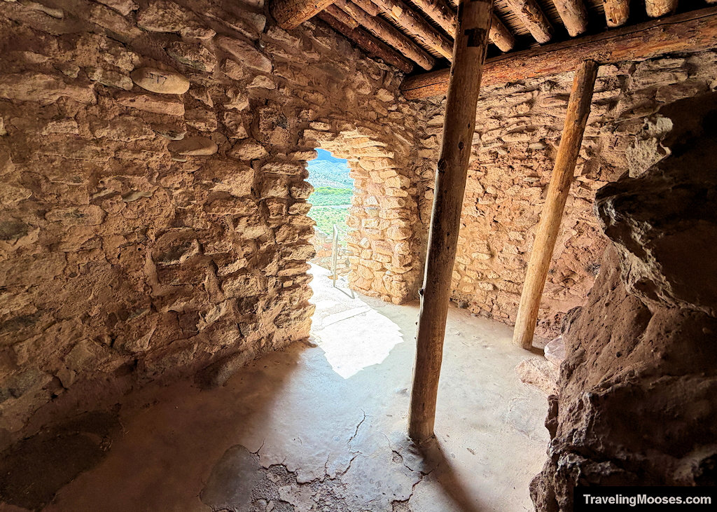 Interior stone room showing upright wooden support posts, rough ceiling logs, and an arched stone doorway opening to a sunny valley view