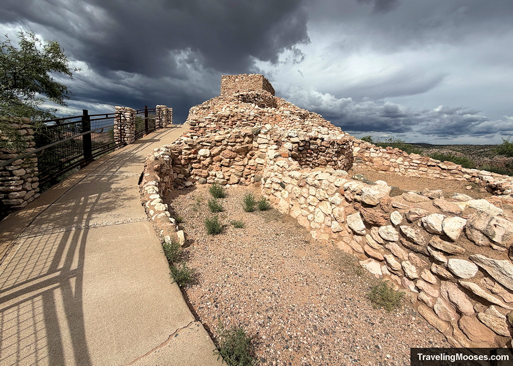 Paved sidewalk curving past low stone walls toward the square citadel tower under an ominous dark storm sky