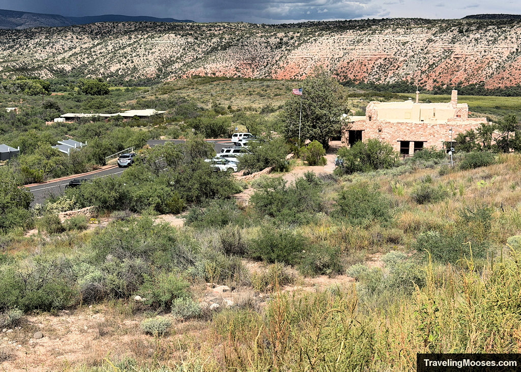Elevated overlook of the stone visitor center building, parking area, and desert valley hills from the ruin ridge