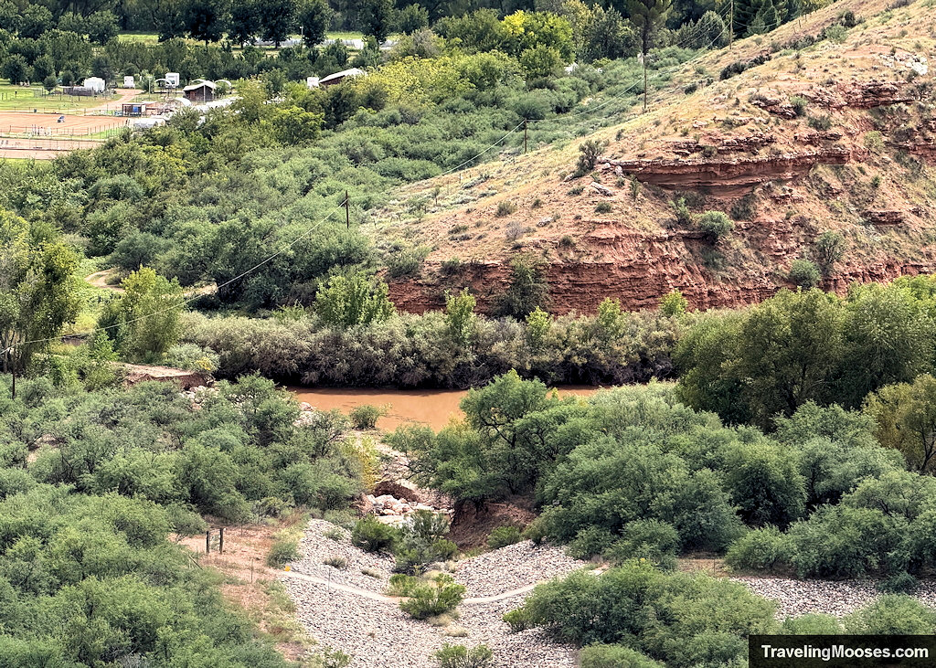 Telephoto view of a brown desert stream framed by lush green trees, red rock bluffs, and a dry gravel wash