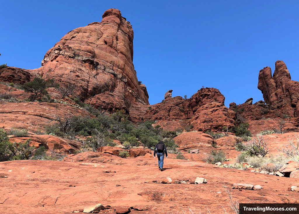Solo hiker walking up a wide expanse of open red slickrock toward a massive rock formation