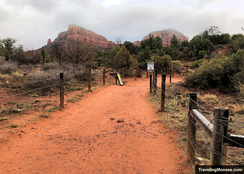 Red dirt trail entrance flanked by wooden post and wire fencing heading toward distant sandstone buttes
