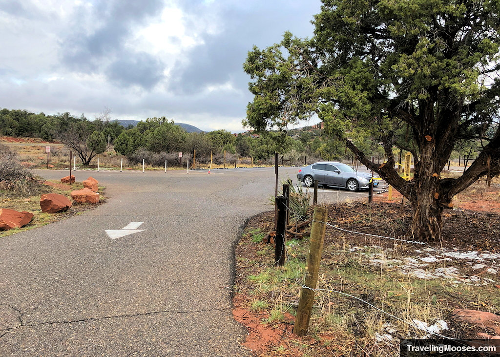 Paved trailhead parking lot with single parked car next to a juniper tree and light morning snow dusting