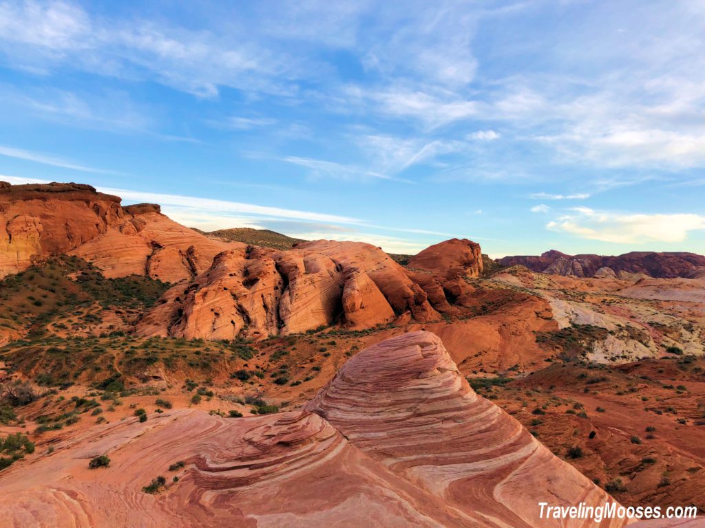 The famous Fire Wave rock formation with red and white sandstone layers against a back-drop of more red sandstone rock formations and blue skies.