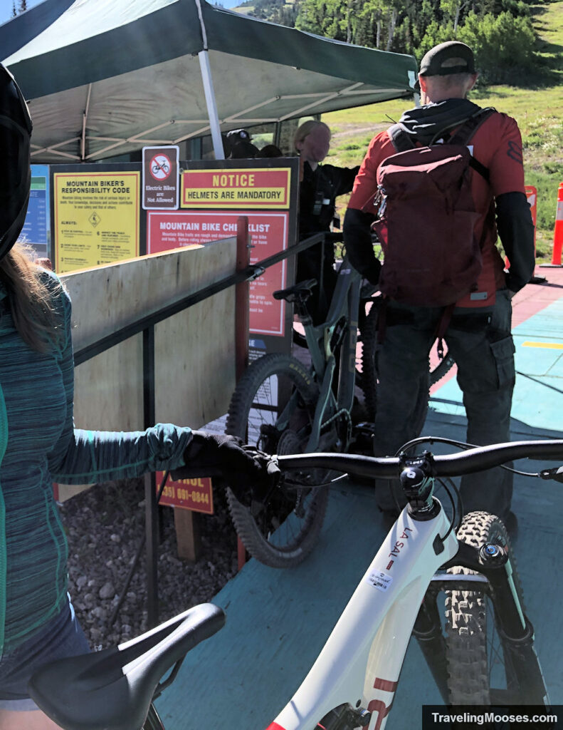 Riders waiting to load mountain bikes onto the Giant Steps lift at Brian Head, with safety signs visible at the entrance.
