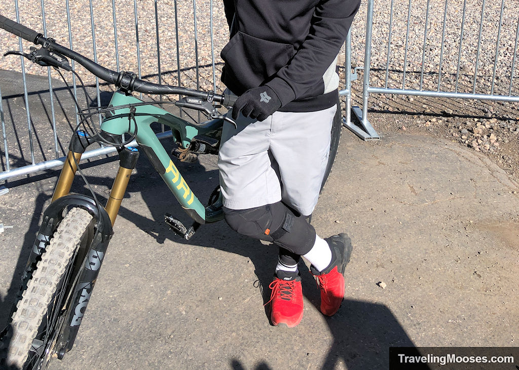 Rider standing with a Brian Head rental enduro bike, wearing gloves and knee pads before starting the Color Flow trail.
