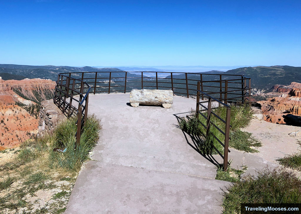 Railed viewing platform and log bench at the Sunset View Overlook in Cedar Breaks, with distant cliffs in the background.