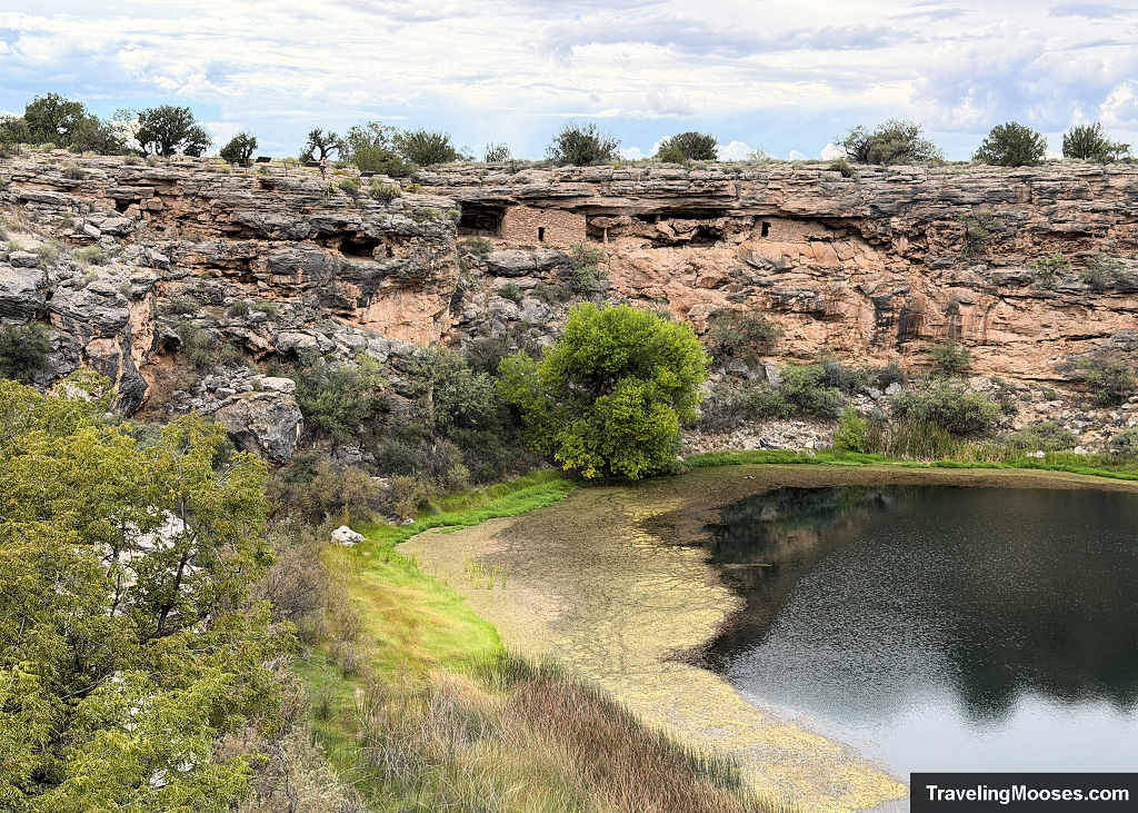 Elevated view across Montezuma Well sinkhole showing ancient Sinagua cliff dwellings built into the limestone rock face above the deep green spring water.