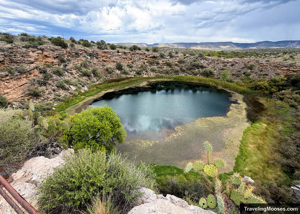 Wide overlook view of the deep green limestone sinkhole at Montezuma Well National Monument under a dramatic, cloudy sky.